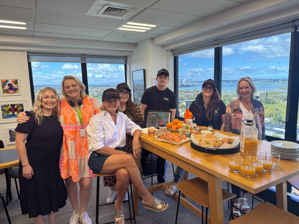 Seven people gather around a table with bagels, fruit, juice, and snacks in a bright office with city views, celebrating as PD turns five.