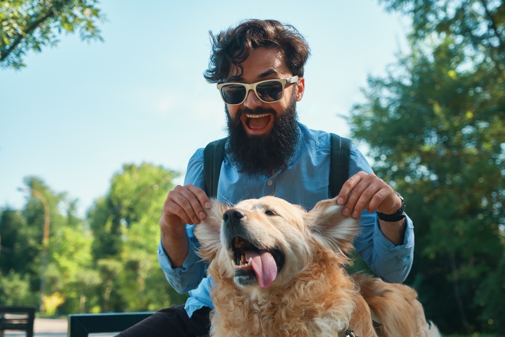 A man with a beard and sunglasses playfully holds up a golden retriever's ears outdoors; both appear happy and relaxed—perfect inspiration for gifts for pet lovers or a unique cat dad Fathers Day gift.