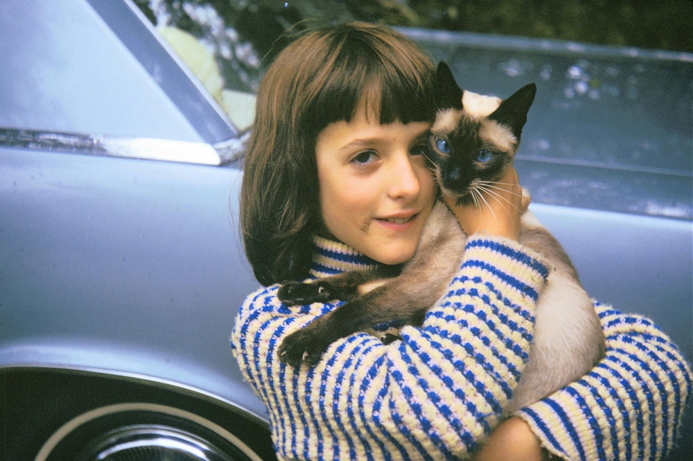 A young girl in a striped sweater, showing what makes PD different, holds a Siamese cat in her arms while standing next to a blue car.