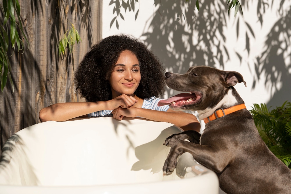 A woman smiles while sitting in an empty white bathtub outdoors, as a happy dog with a bright orange collar—showing what makes PD different—rests its front paws on the tub's edge.