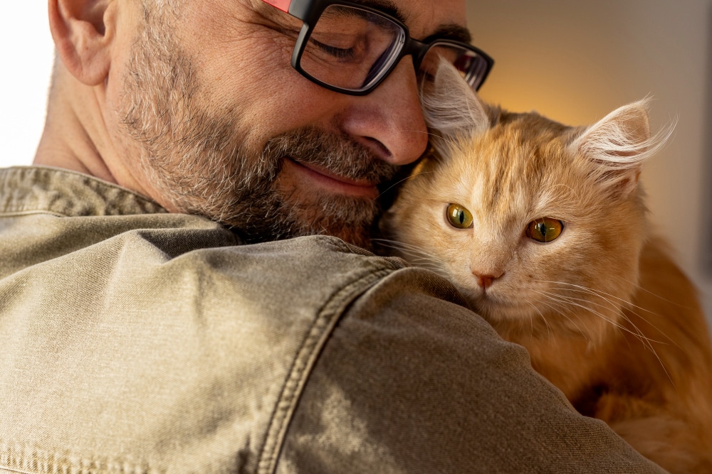 A man with glasses and a beard smiles while holding an orange cat on his shoulder.