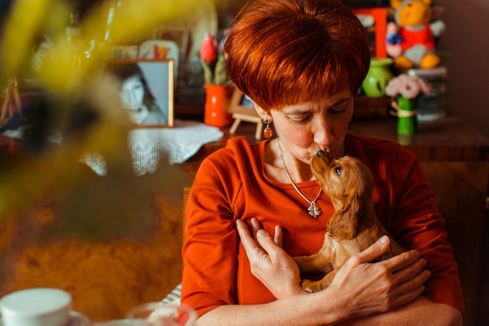 A woman with short red hair holds a small brown puppy and kisses its nose in a cozy, decorated room, framed photos and toys highlighting what makes PD different—warmth, connection, and loving moments.