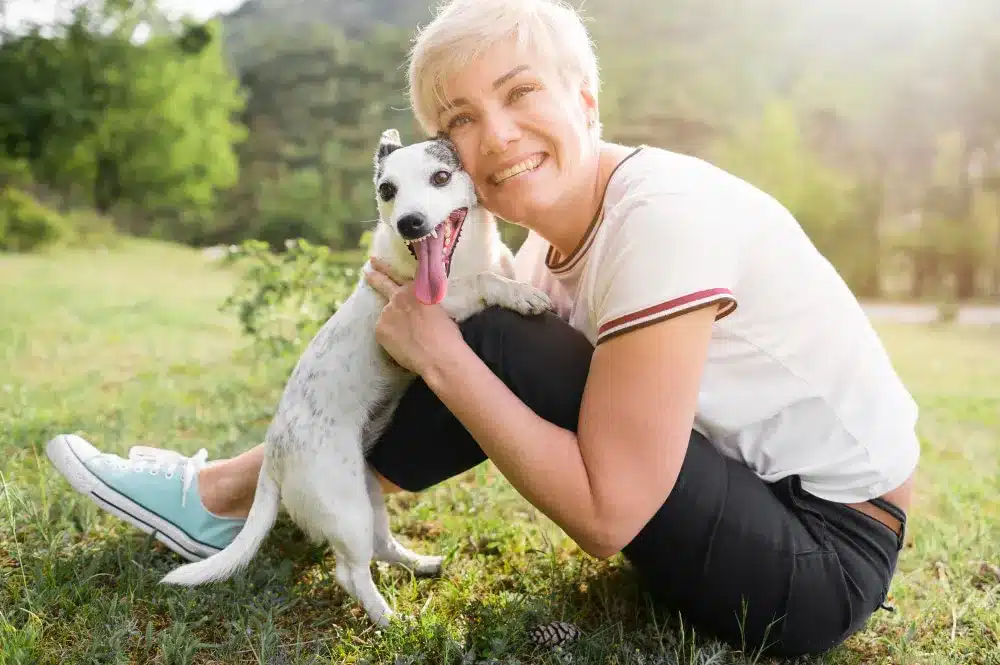 A person crouches on grass outdoors, smiling and hugging a small white and black dog with its tongue out, capturing the joy found in the human-pet bond survey.