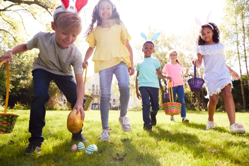 Five children wearing bunny ears participate in an Easter egg hunt outside, each holding a basket and searching for colorful eggs on the grass, while an Easter dog watches playfully nearby.