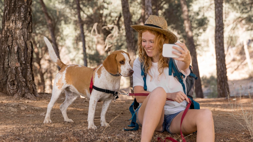 A proud dog mum wearing a hat sits on the ground in a forest, holding a phone and taking a selfie with her brown and white dog on a leash.