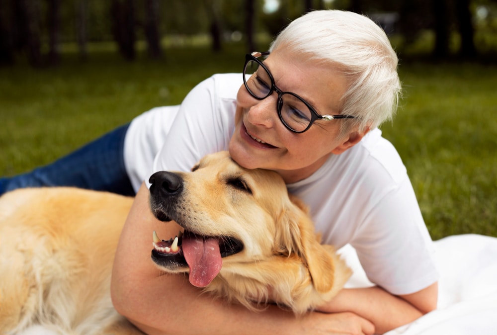 Smiling pet mum with short gray hair and glasses lies on grass, hugging her happy golden retriever outdoors.