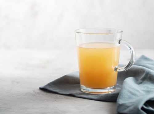 A clear glass mug filled with orange-colored juice sits on a light blue cloth, while a curious cat watches from the side against a neutral background.