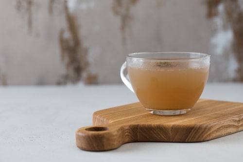 A glass cup filled with light brown bone broth recipe sits on a wooden cutting board against a neutral background.