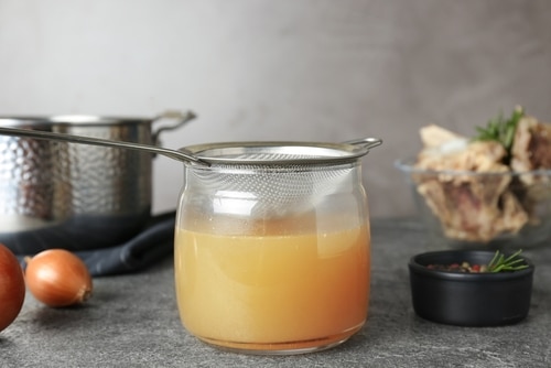 A jar of homemade bone broth is being strained with a metal sieve; onions, bones, a pot, and a small bowl of spices are nearby on a gray countertop—perfect for trying in your next dog-friendly bone broth recipe.
