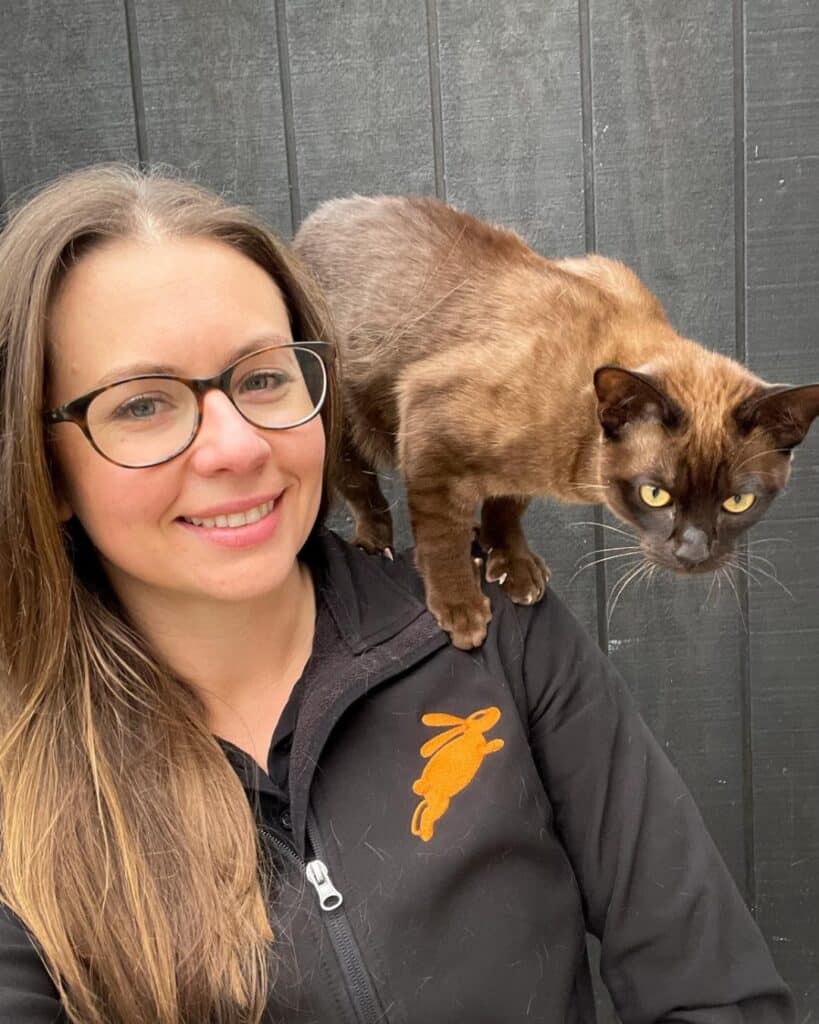 A woman with glasses smiles at the camera while a brown cat stands on her shoulder in front of a black wooden wall, capturing a joyful moment as pd turns 6.