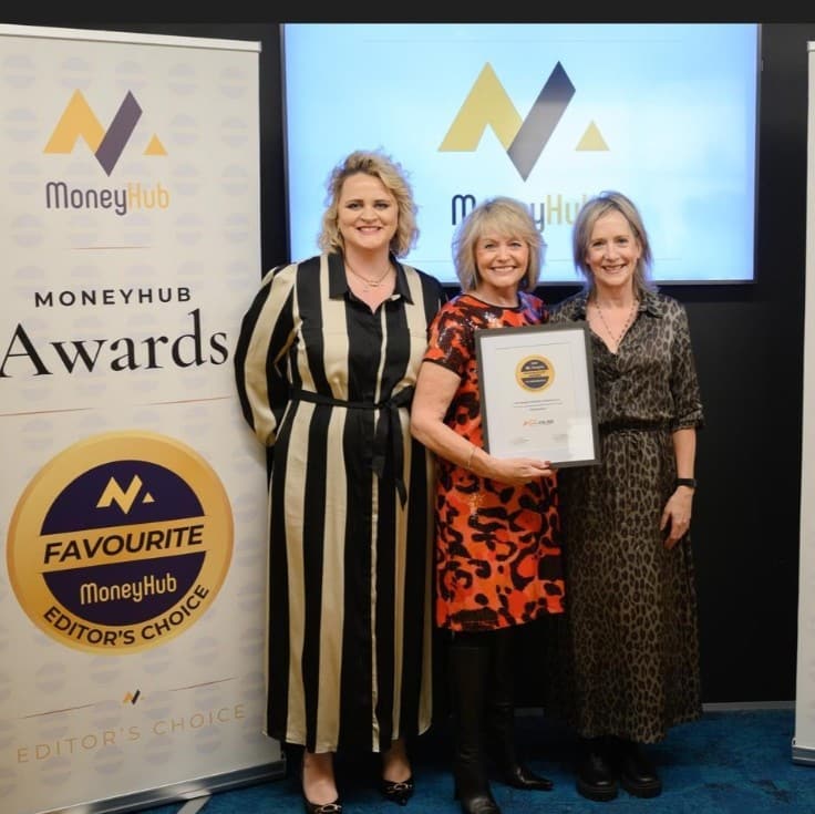 Three women stand smiling at a MoneyHub Awards event, with one holding a framed certificate. Banners displaying “MoneyHub Awards” and “Favourite Editor’s Choice” are visible.