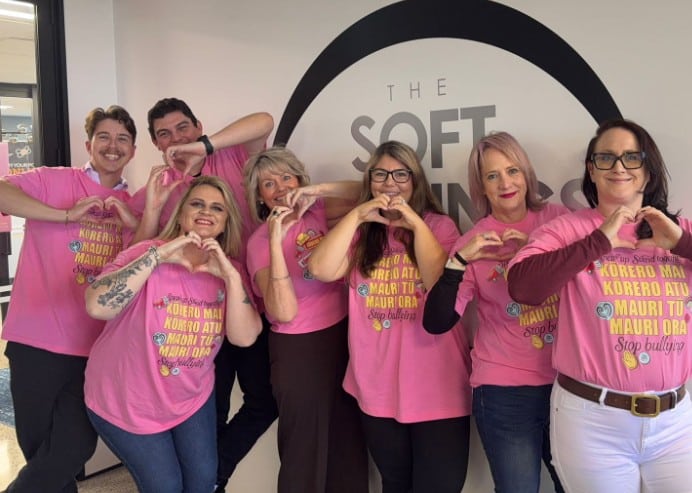 Seven people stand in a row wearing matching pink shirts, smiling, and making heart shapes with their hands in front of a wall sign that reads “THE SOFT”.