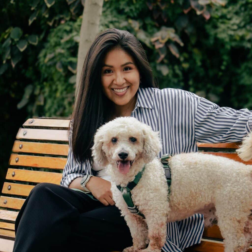 A woman in a striped shirt sits on a wooden bench with a small, white curly-haired dog in a green harness, highlighting the mental health benefits of pets.